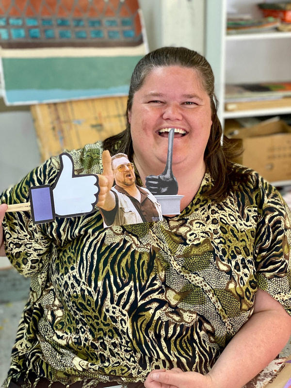 Photograph of artist Kayla Hood, laughing and holding three sticks with thumbs up images