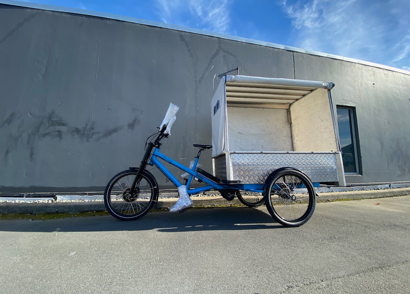a photograph of a three wheel trike next a wall, the trike is an electric cargo bike. Clicking the image takes you to a web page with information about the summer workshops held in January 2022